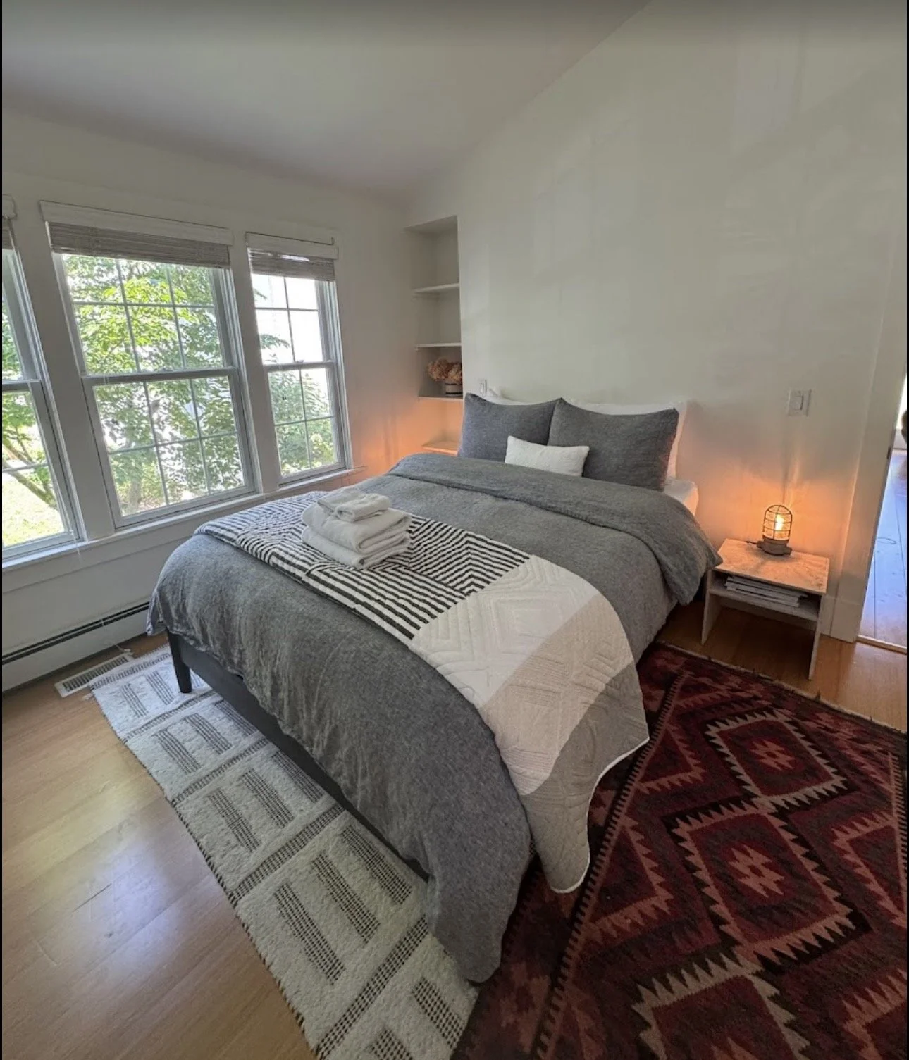 A cozy bedroom with a bed featuring gray and white bedding, flanked by two windows with trees outside, a nightstand with a lamp, and a patterned rug on a wooden floor.
