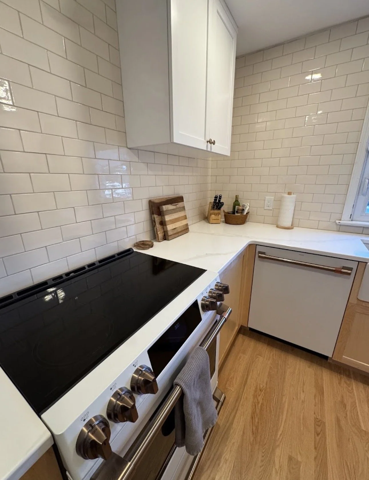 Kitchen with cream-colored subway tile backsplash, white countertops, and wood cabinets. A black stovetop, a towel hanging from the oven handle, and various small kitchen items on the counter, including a cutting board, knife block, and paper towel roll.