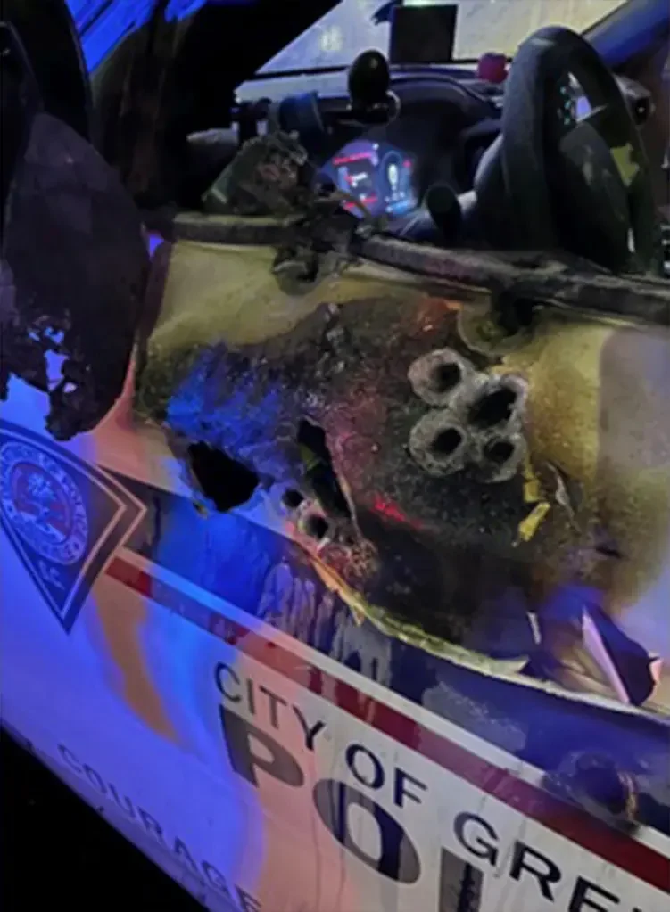 Post offcier ambushed vehicle Close-up of burnt police car dashboard and windshield with a damaged front section, showing entry holes, with a police badge and city of Greenville South Carolina sign partially visible.
