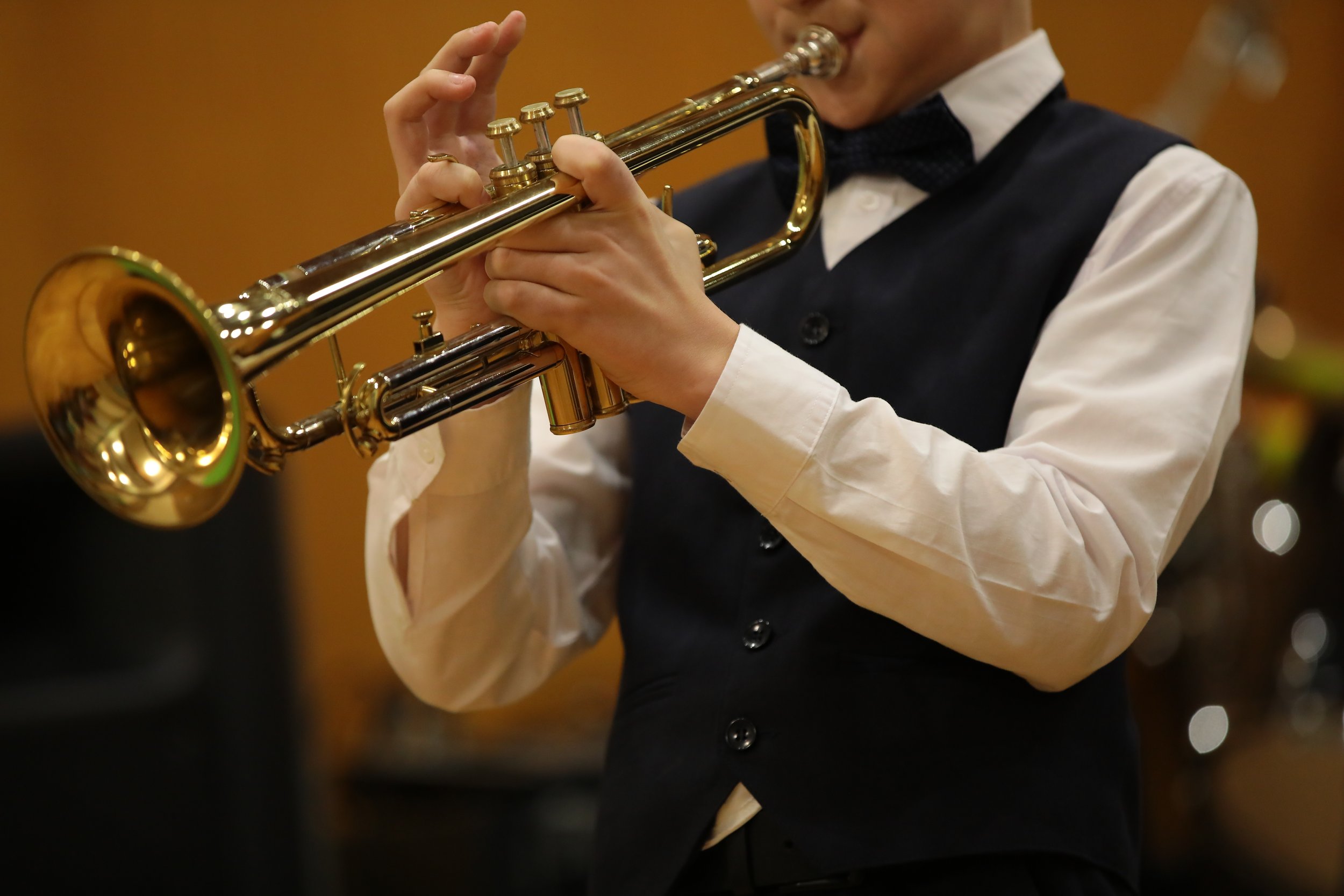 A person wearing formal attire, including a white shirt, black vest, and bow tie, playing a gold trumpet.