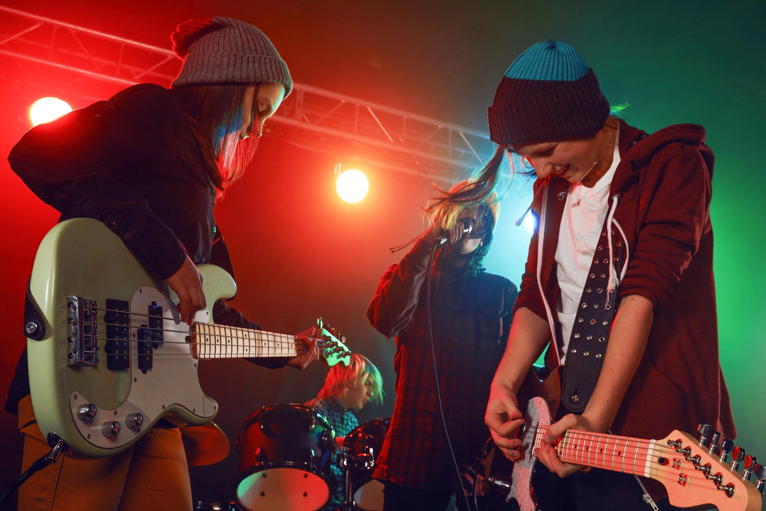 A group of young musicians performing on stage; three females with guitars and a drummer, under vibrant stage lighting.