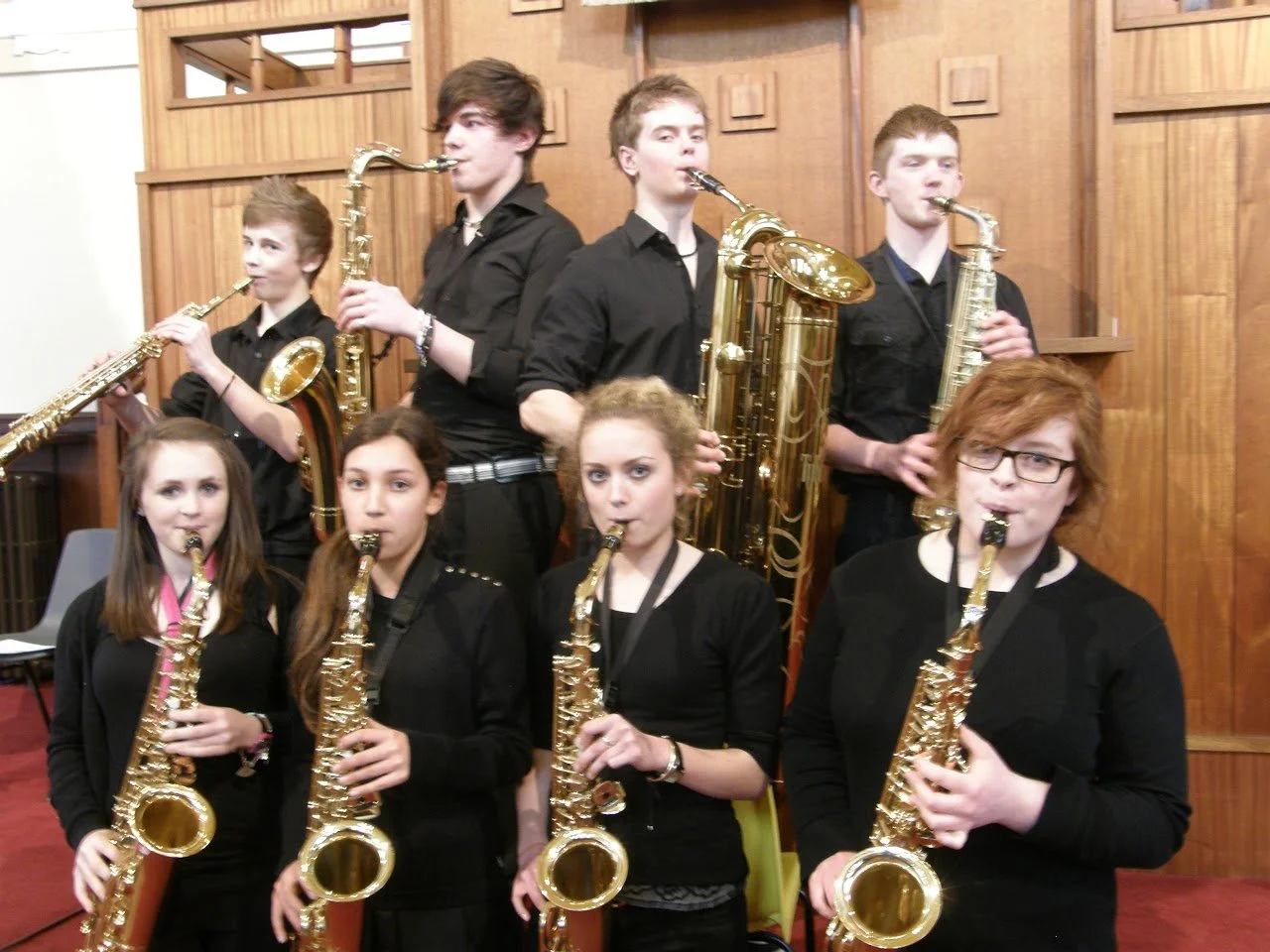 Seven young musicians, six females and one male, playing saxophones in a wooden room with paneling.