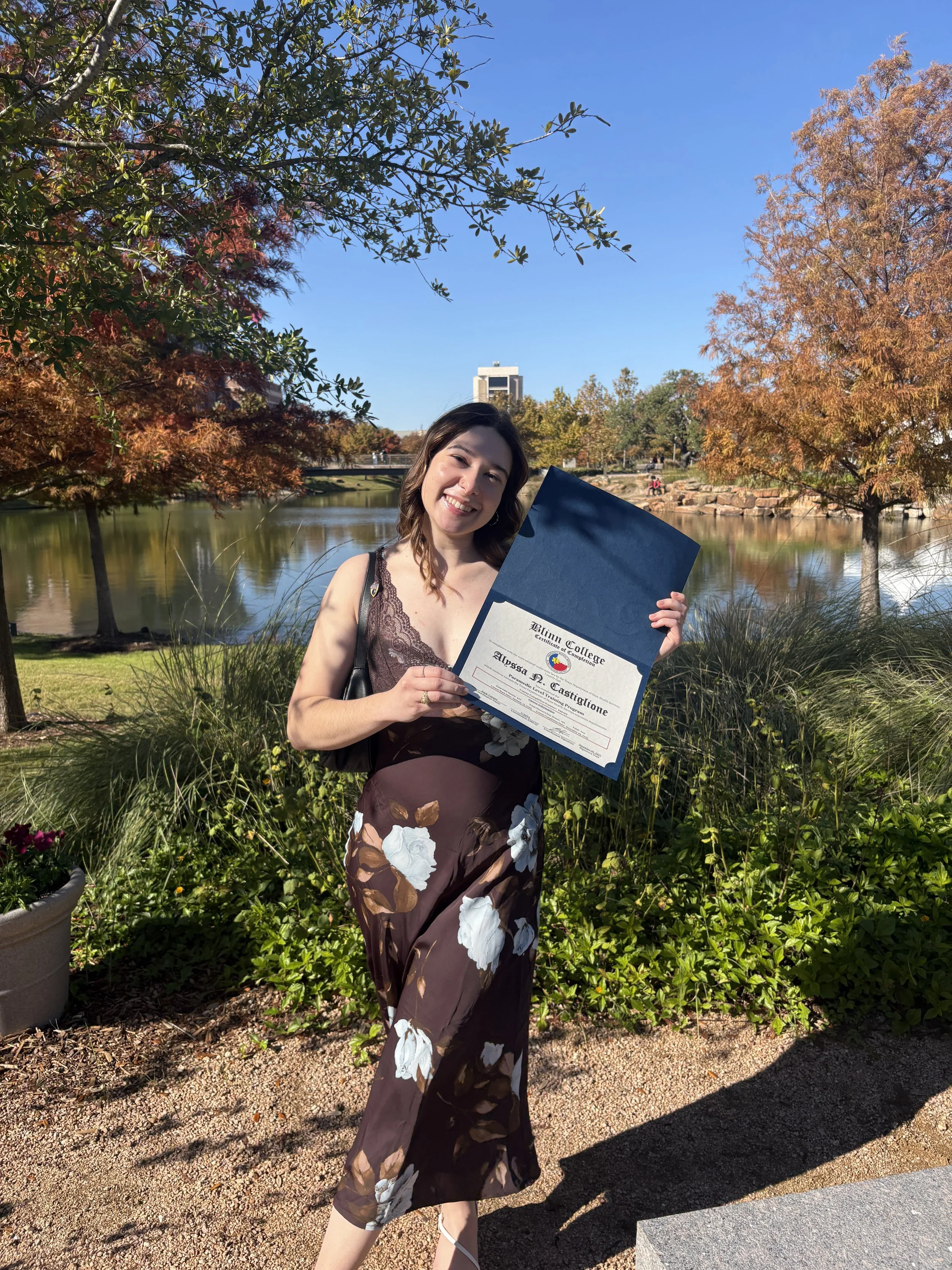 A young woman smiling and holding a diploma outdoors on a sunny day, standing on a pathway near a pond with trees in fall colors.