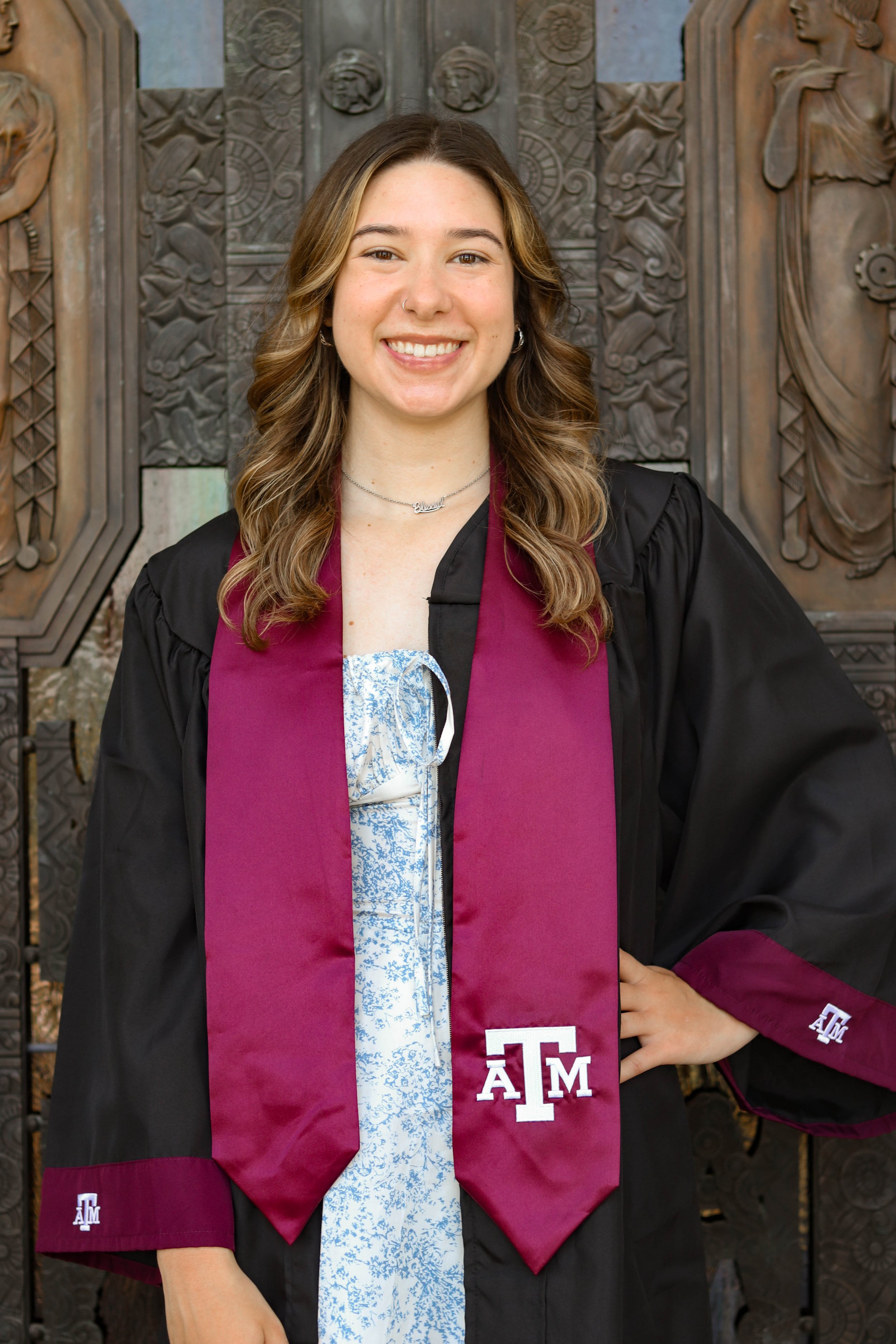 A young woman in graduation attire, wearing a black gown with maroon accents and a maroon stole with white Texas A&M logos, standing in front of a carved wooden background, smiling with hand on her hip.