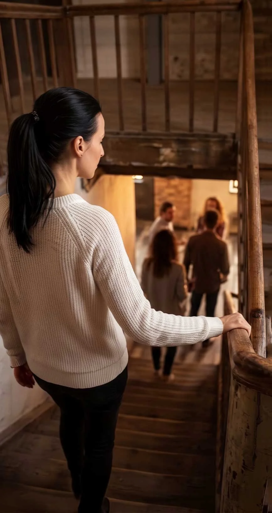 Eine Frau mit dunklem Haar in einem weißen Pullover, die auf einer Holztreppe steht und nach unten blickt, wo eine Gruppe von Leuten in einem rustikalen Raum miteinander spricht.