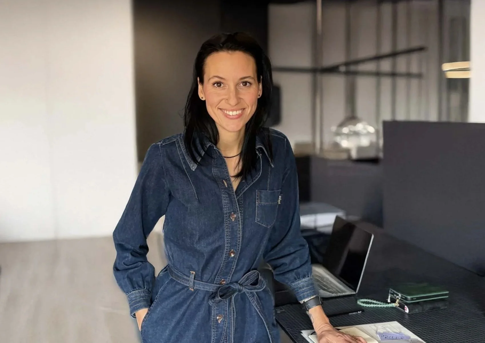 Eine lachende Frau mit schwarzen Haaren, die eine Jeans-Kleid trägt, steht an einem Schreibtisch mit einem Laptop und Papieren in einem modernen Büro.