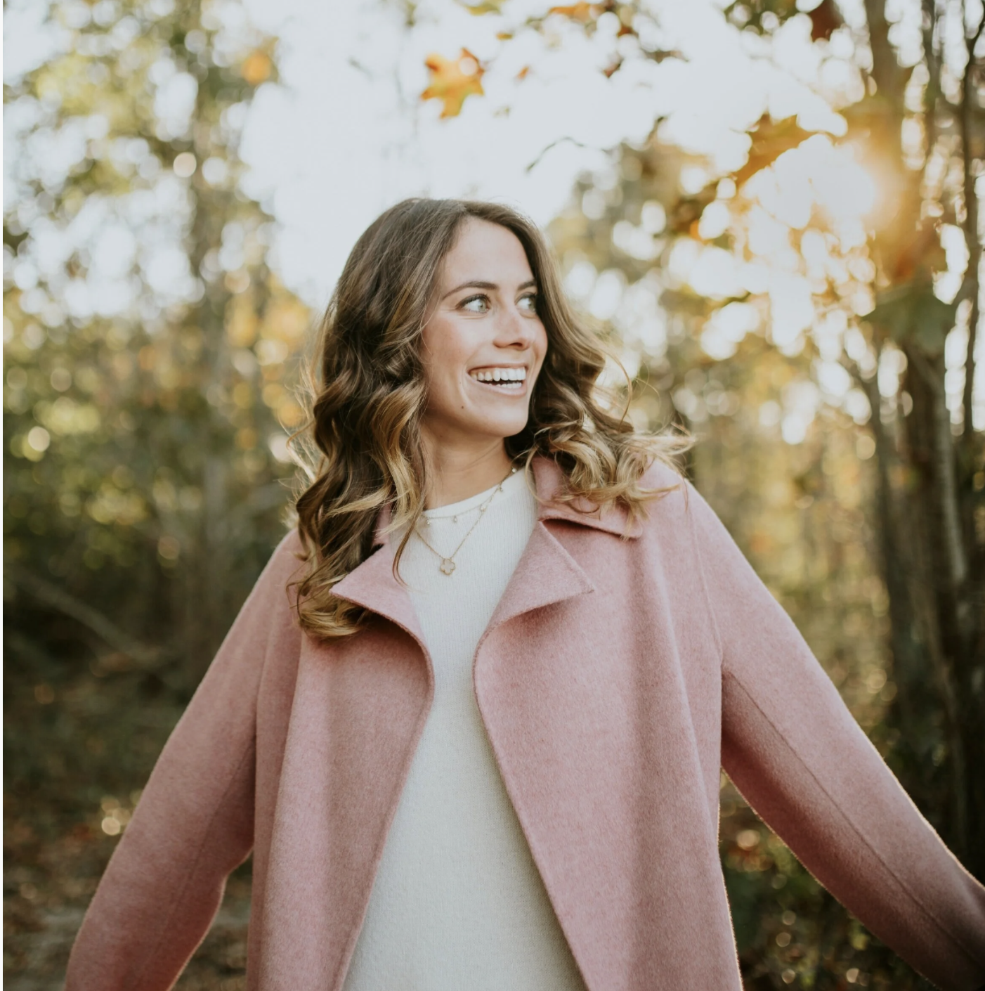 A woman with curly brown hair smiling outdoors in a forested area during autumn, wearing a pink coat over a white top.