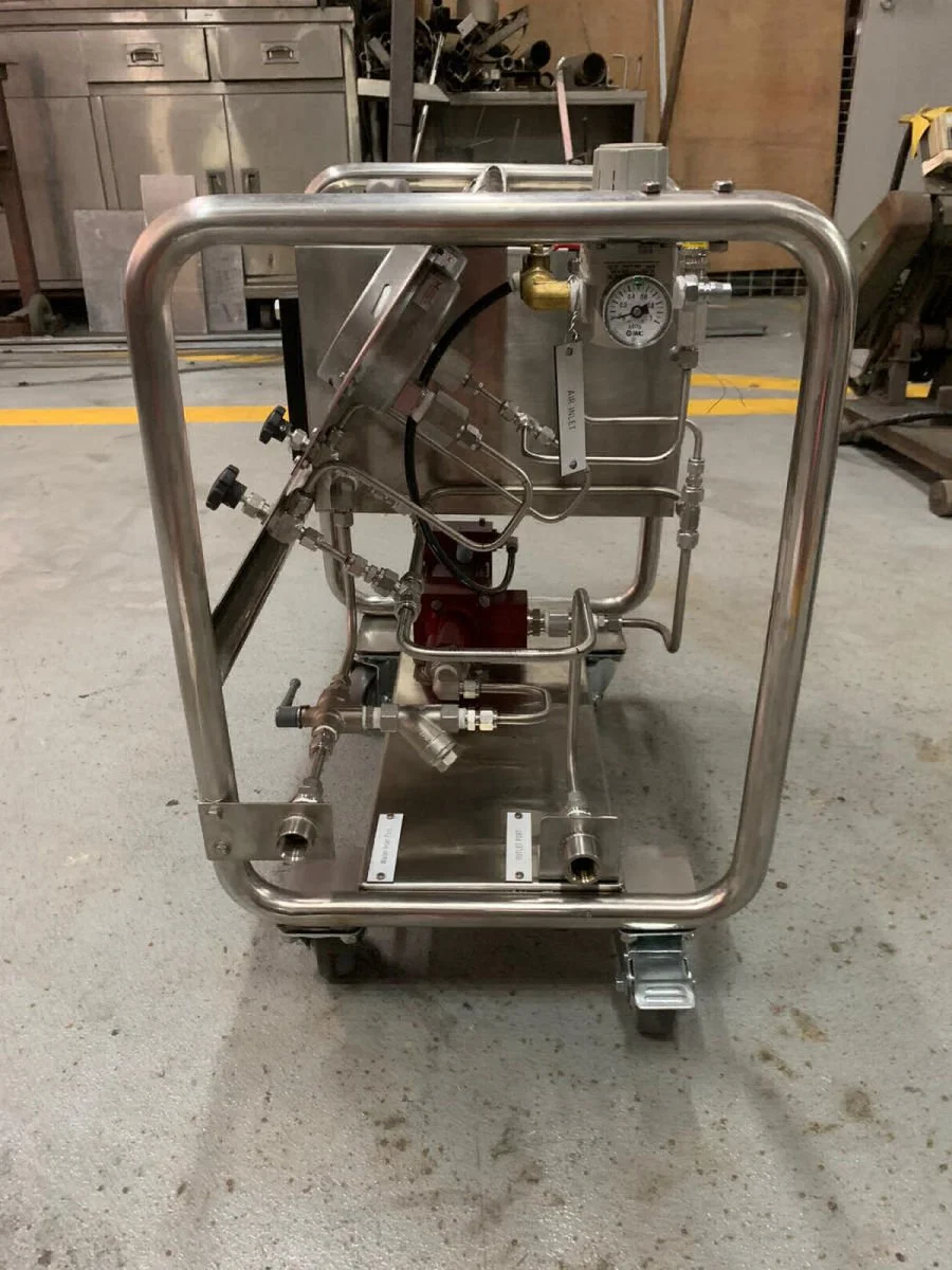 Stainless steel industrial equipment with pipes, a gauge, and valves on a metal cart in a workshop or factory setting.