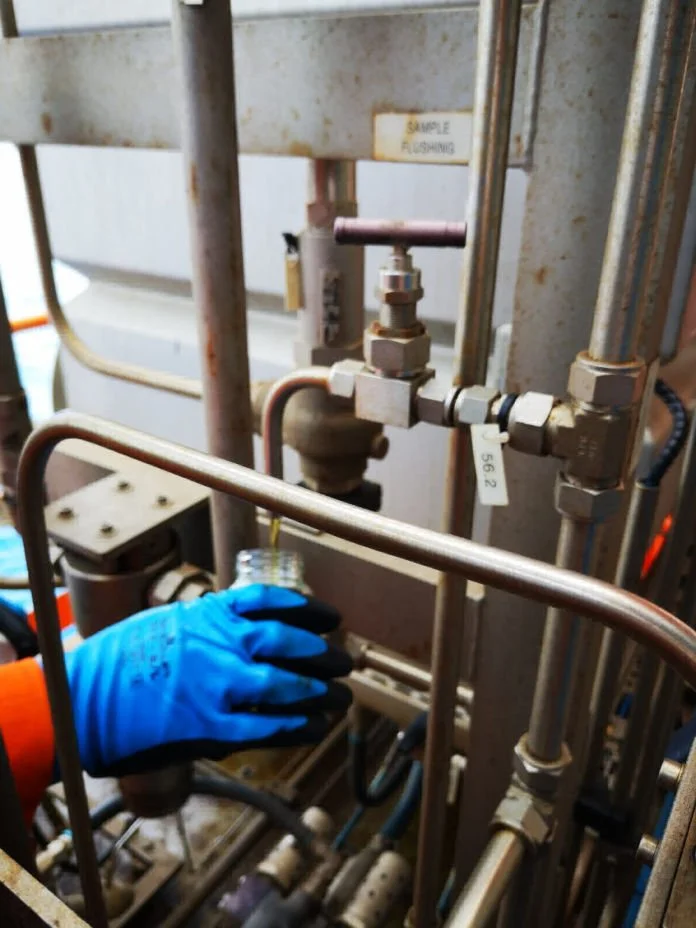 A person wearing blue gloves handling a glass container in an industrial setting with stainless steel pipes and valves.