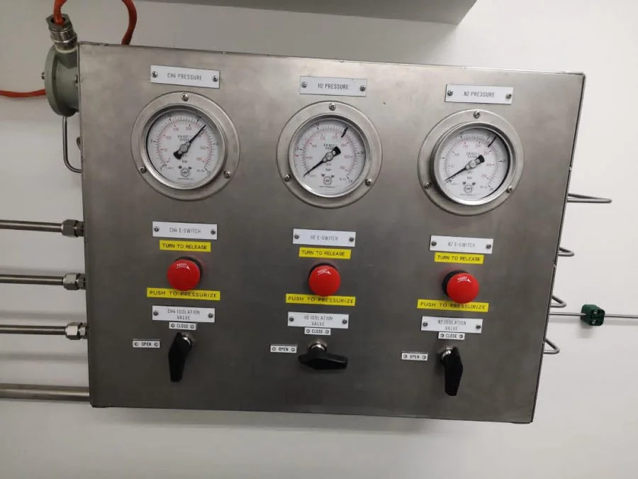 Metal control panel with three pressure gauges, red emergency stops, black switches, and yellow labels with instructions, mounted on a wall with pipes.