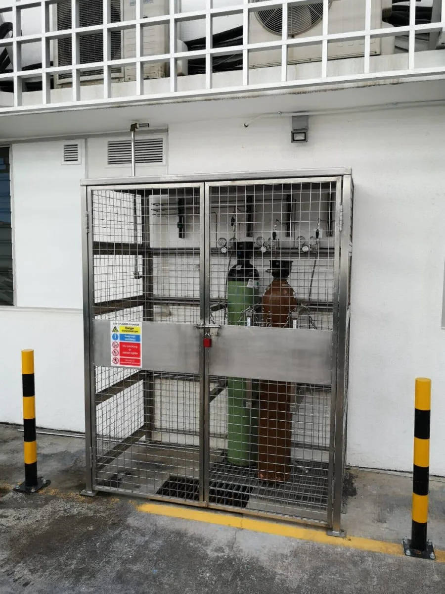 A fenced storage enclosure containing gas cylinders in front of a white building, with air conditioning units and balconies visible above.