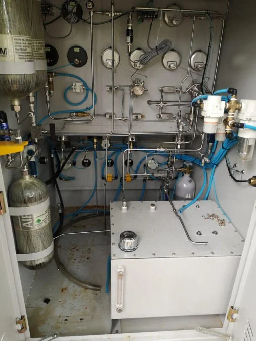 This image shows a laboratory instrument with various tubes, valves, and regulators, including a large gas cylinder on the left and a white control unit with blue tubing on the right.