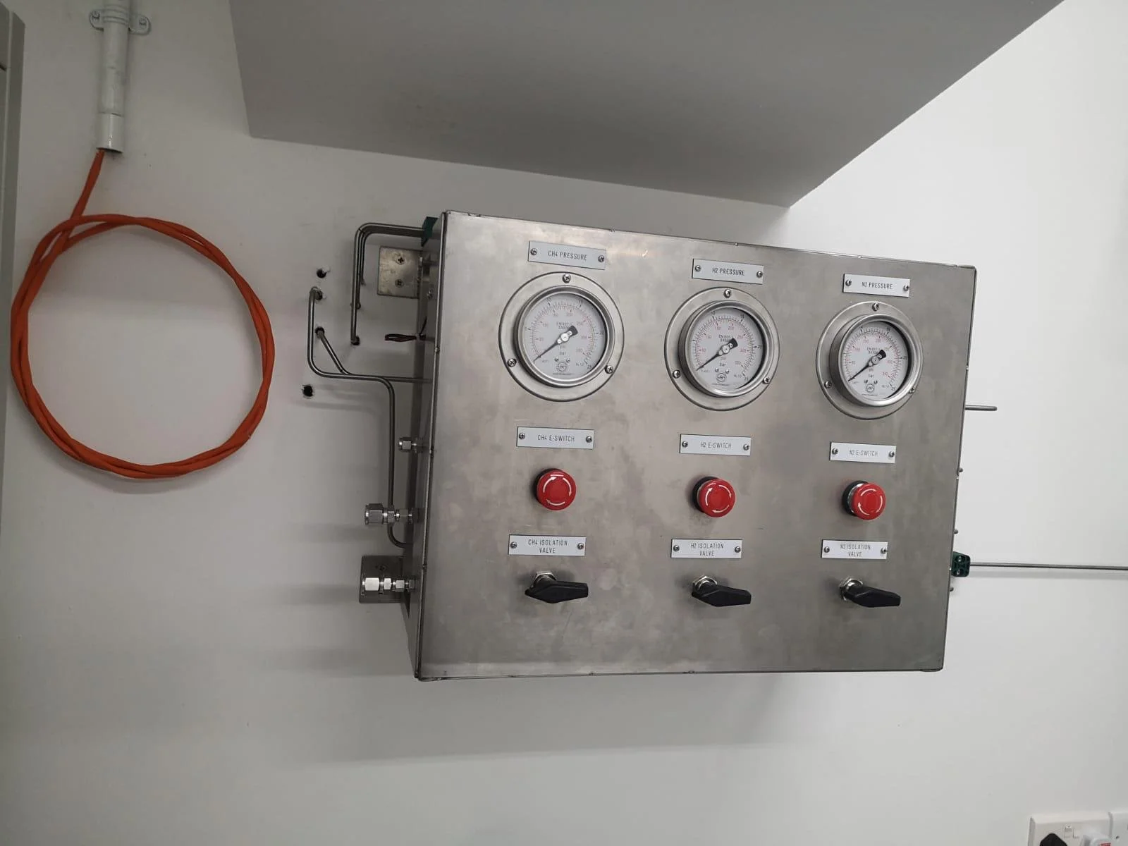 A metal control panel with three pressure gauges labeled CH1 PRESSURE, CH2 PRESSURE, and CH3 PRESSURE, each with a black knob below them, connected to various pipes and wires, mounted on a white wall.
