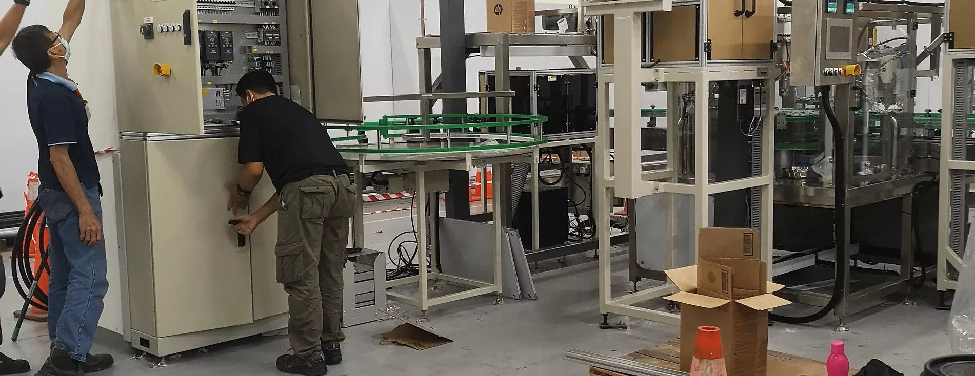 Two workers in a factory or industrial setting working on electrical or machinery panels surrounded by equipment and tools.