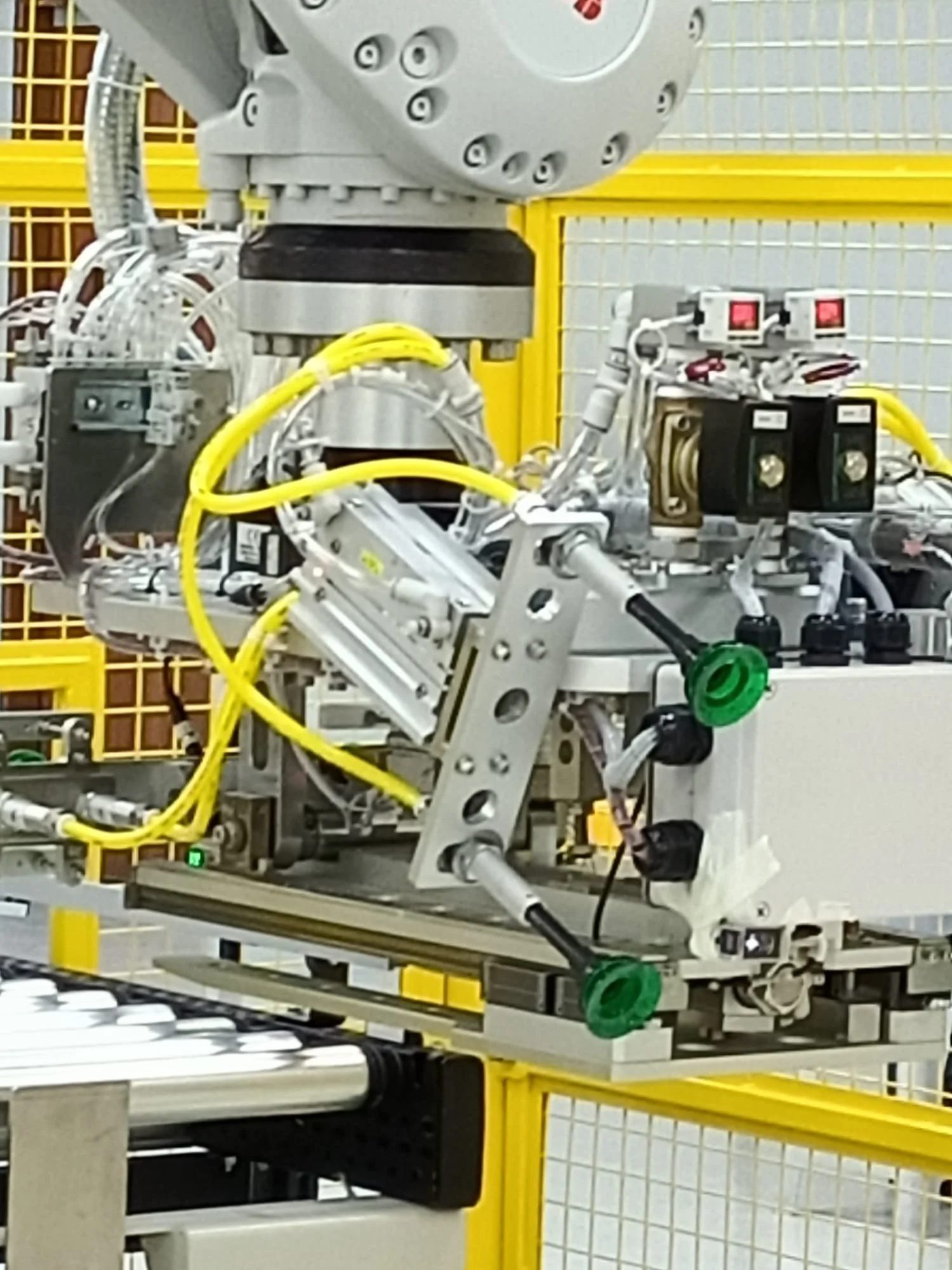 Close-up of a complex industrial robotic arm with various wires and components, set in a manufacturing or assembly environment with yellow safety barriers in the background.