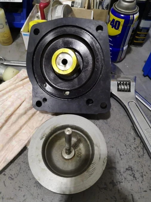 Disassembled mechanical part on workbench, with a black part with a yellow center and a separate silver metallic component with a central pin.