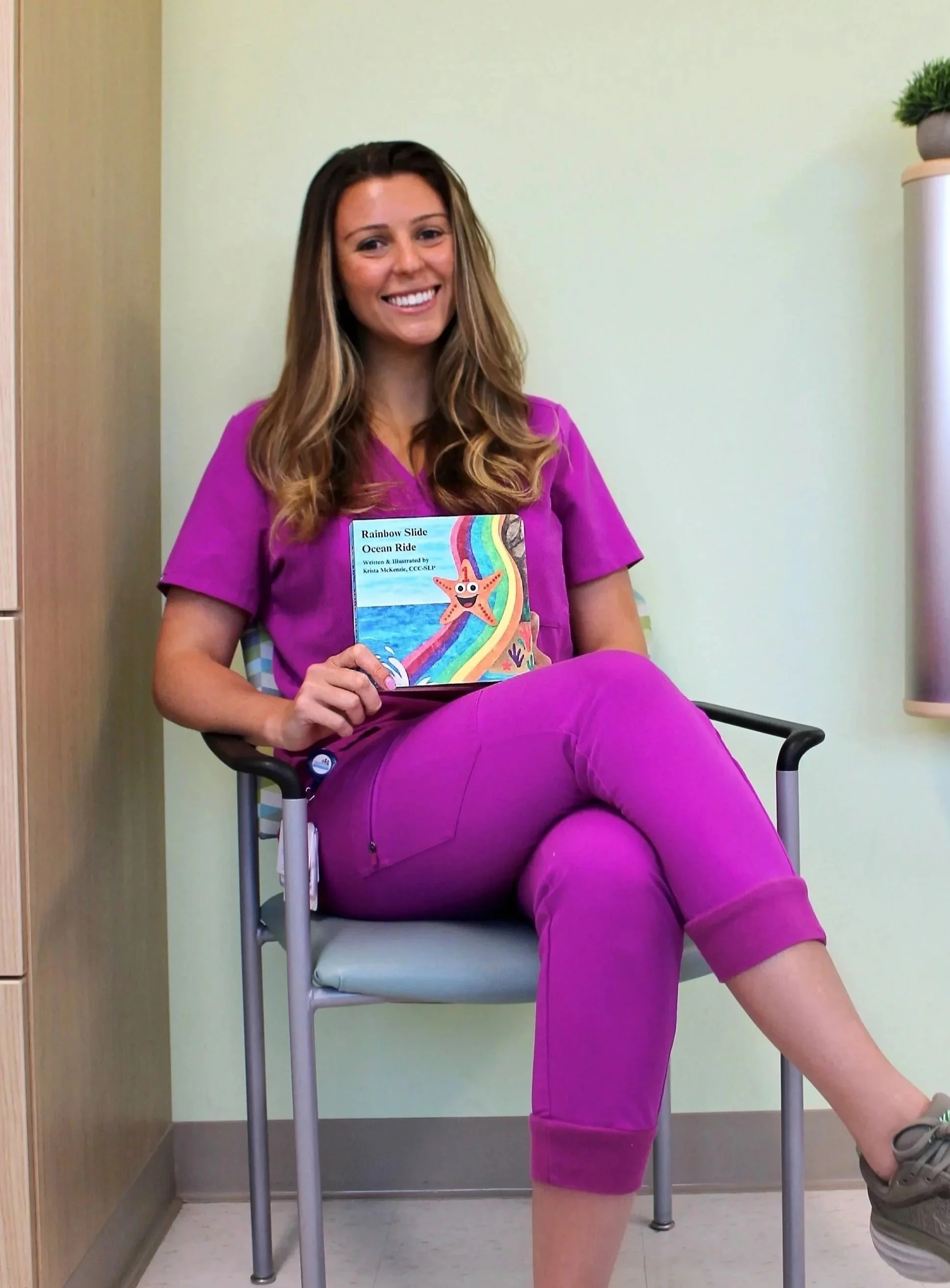 A woman in purple scrubs sitting on a chair holding a children’s book titled 'Rainbow Slide Ocean Ride,' in a medical office or clinic.