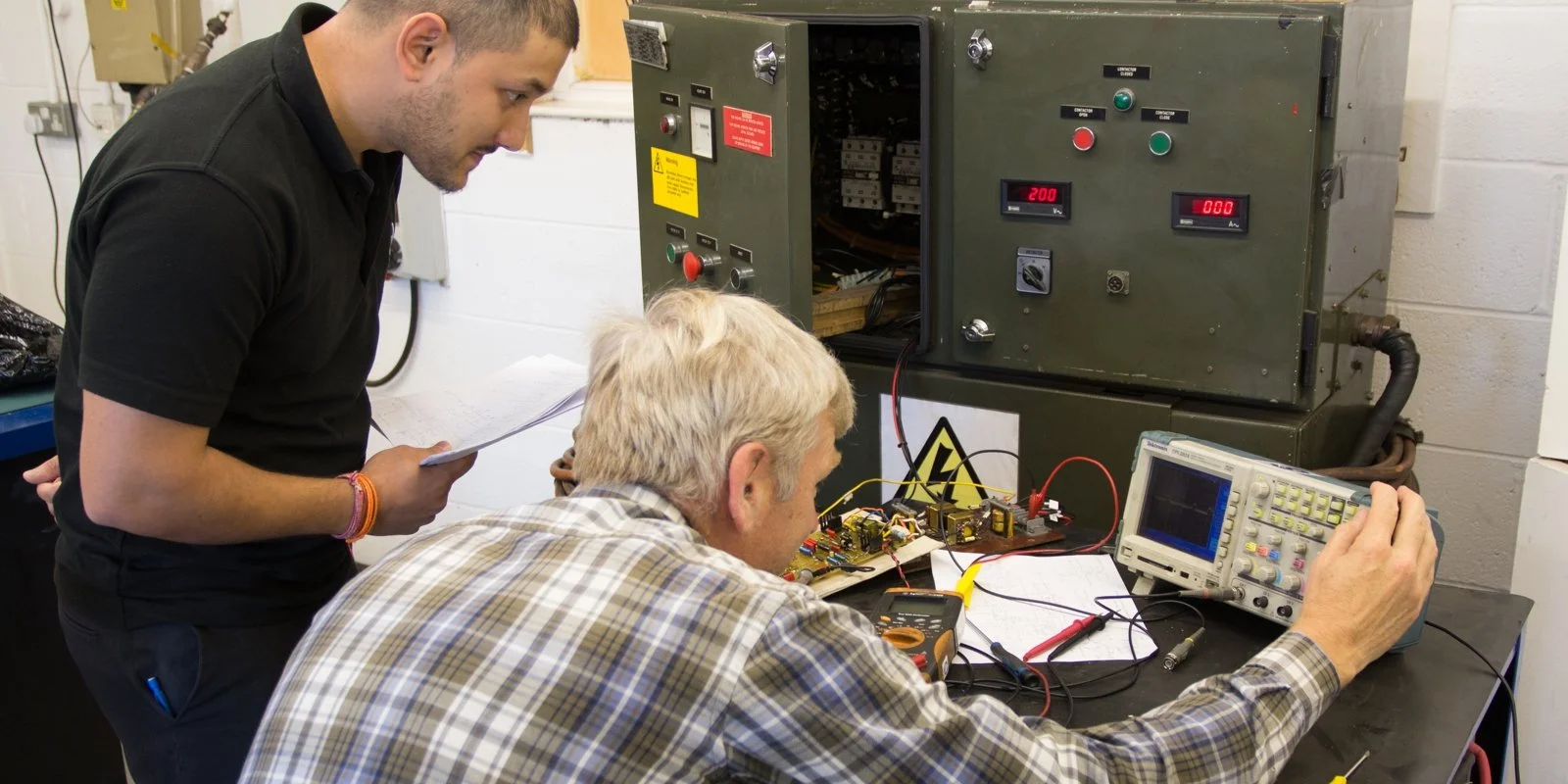 Two men working on an electrical engineering experiment with circuit boards, an oscilloscope, and a high voltage power supply in a lab.