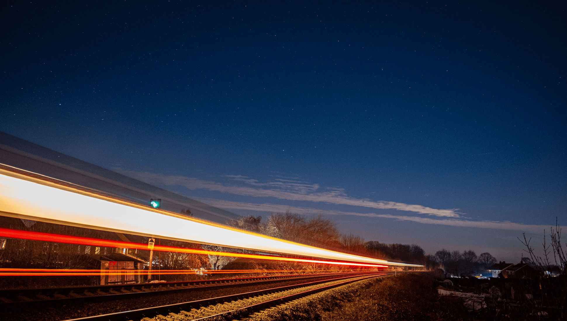 Nighttime scene of a train passing by, creating light streaks on the tracks under a starry sky. Trees and houses are visible in the distance.