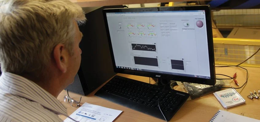 A man sitting at a desk working on a computer with graphs and charts on the screen, with a notebook, pamphlet, and small electronic components on the desk.