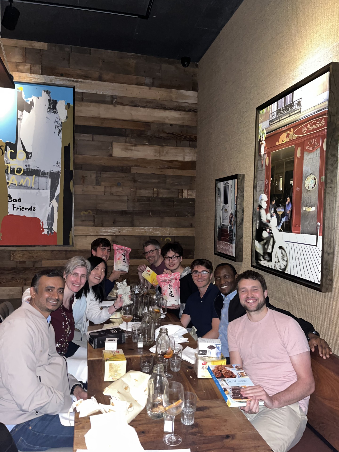 A group of ten people sitting at a long wooden table in a restaurant or cafe, smiling and holding gift boxes.