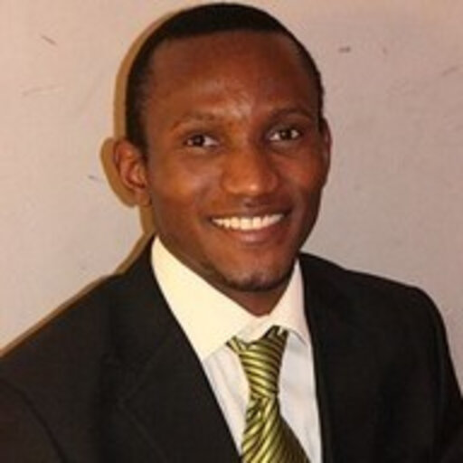 A man wearing a black suit, white shirt, and green striped tie, in front of a plain light-colored wall.