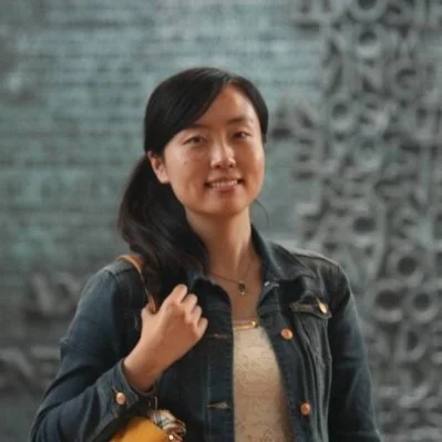 A woman with black hair, wearing a denim jacket, standing in front of an intricate textured gray wall.