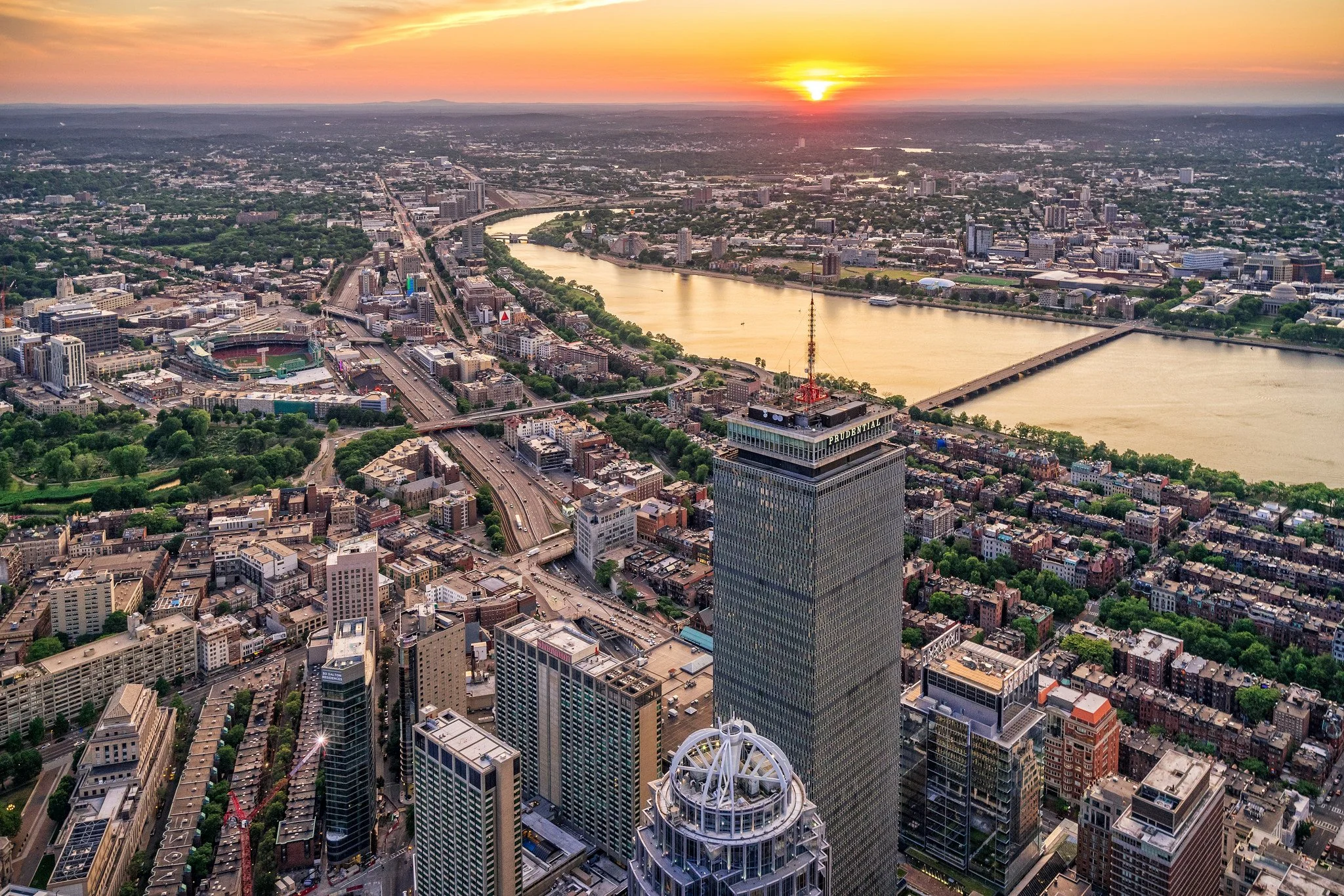 Aerial view of a city skyline at sunset with tall buildings, a river, and bridges.