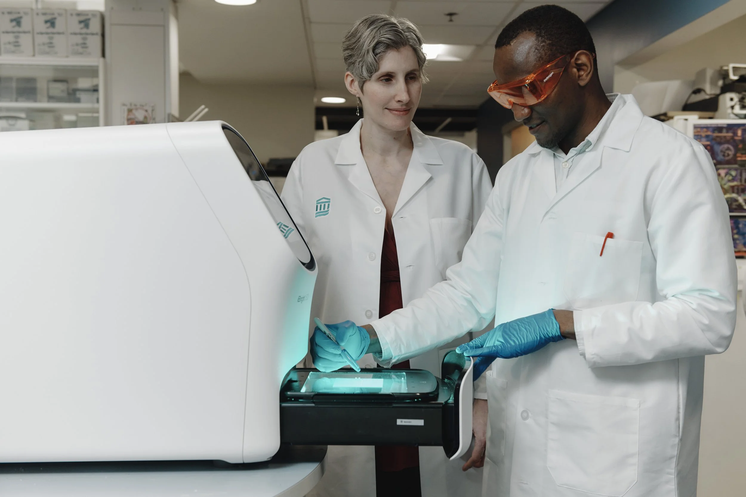 Two scientists in lab coats working with a large laboratory instrument, one is using a stylus on a touchscreen, in a laboratory.