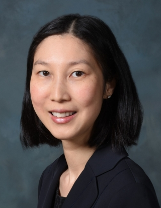 Headshot of a woman with shoulder-length black hair, wearing a dark blazer against a blue background.