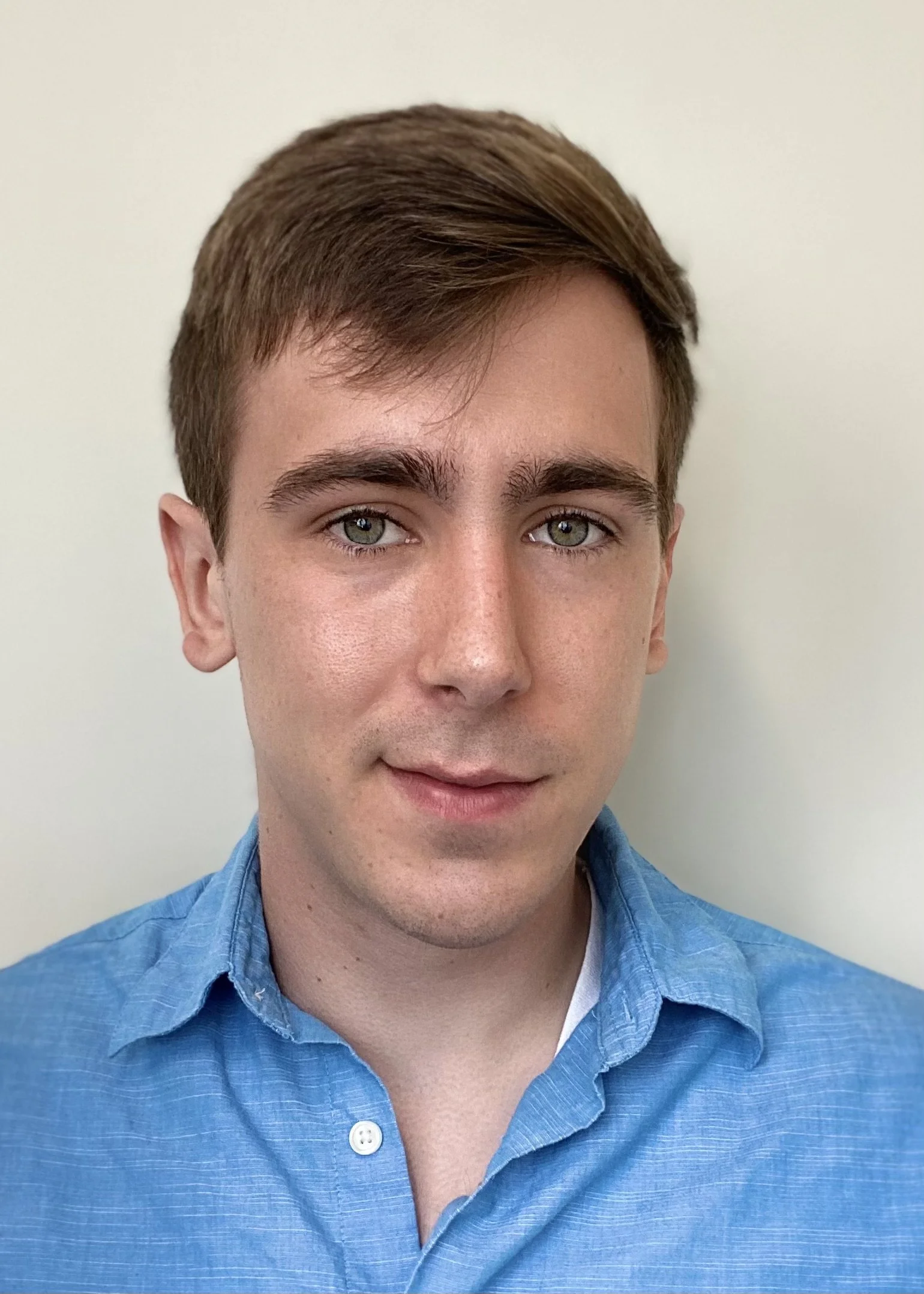 A young man with light brown hair in a blue collared shirt, posing against a plain light-colored background.