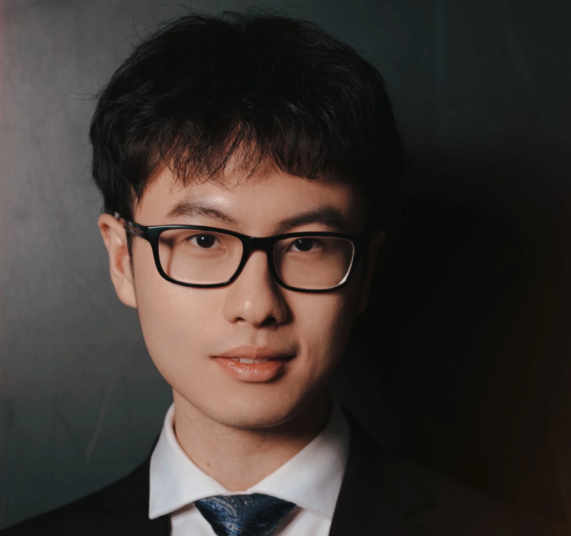 Headshot of a young man with glasses, wearing a suit and tie, against a dark background.