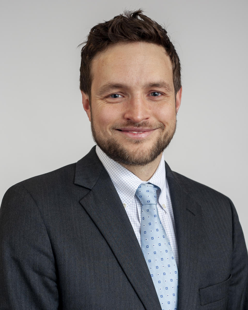 A man with light brown hair and a beard, wearing a dark suit, against a plain light gray background.