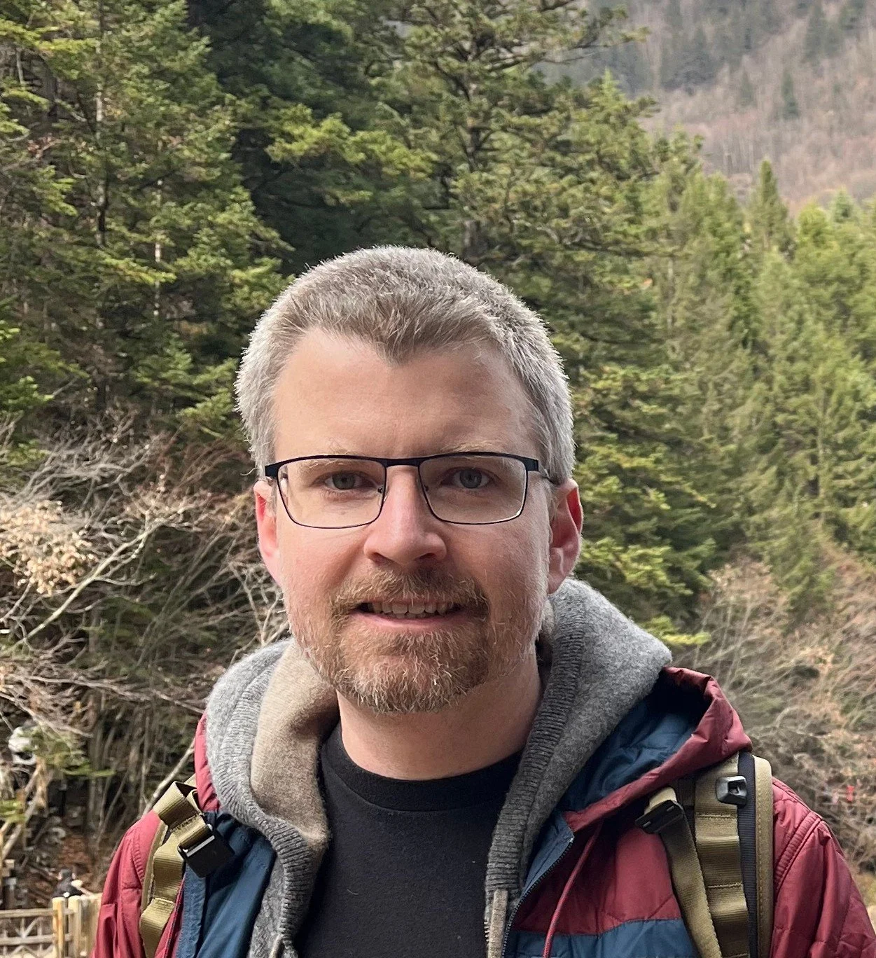 A man with glasses, a beard and short gray hair in front of a forested hillside.
