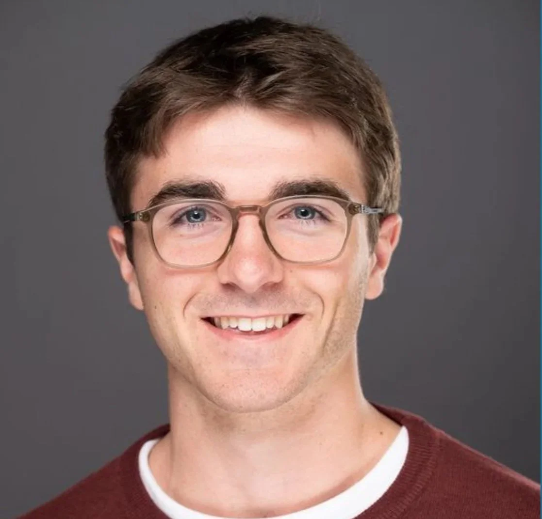Close-up portrait of a young man with brown hair, wearing glasses and a maroon shirt against a gray background.