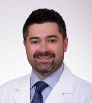 Portrait of a male doctor with dark hair, beard, wearing a white lab coat and blue dress shirt, smiling against a plain background.