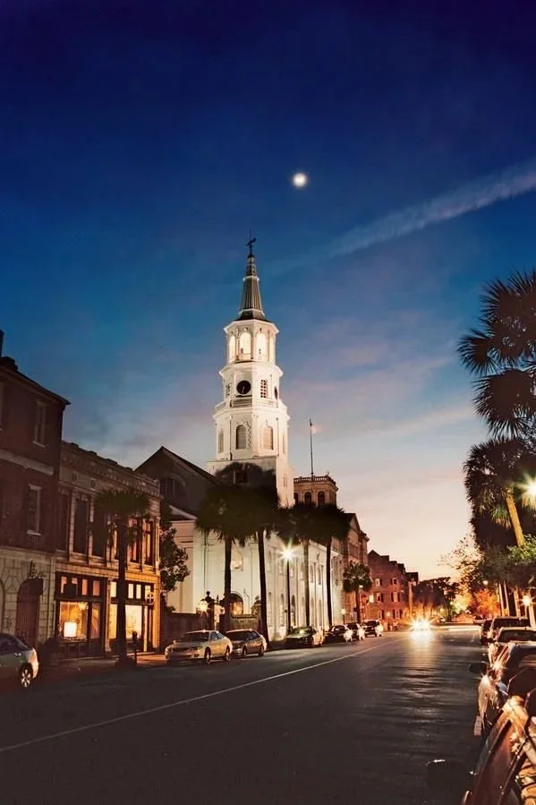 A historic church with a tall spire and clock tower, surrounded by storefronts, parked cars, palm trees, and a clear evening sky with visible moon and clouds.
