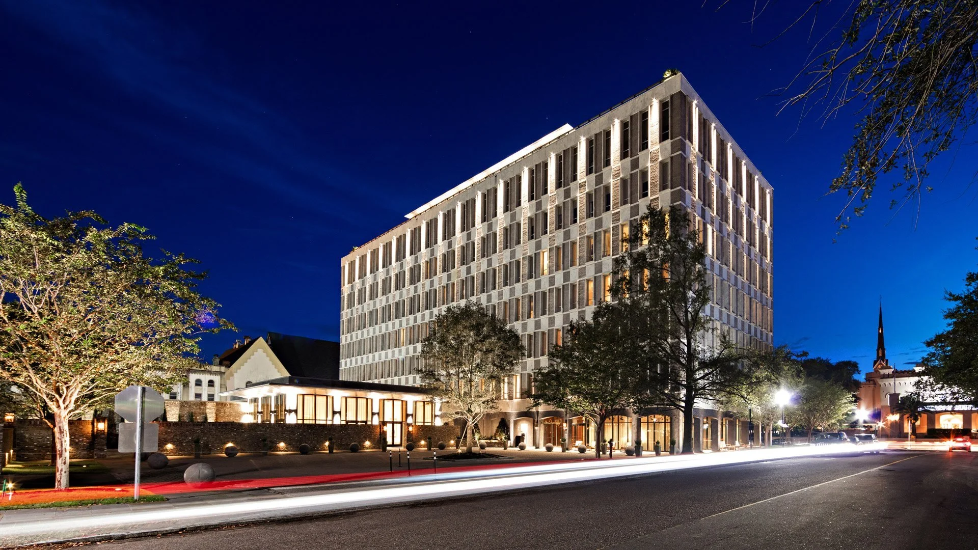 A modern multi-story building illuminated at night with lighted windows, situated beside a street with streaks of car lights from long exposure, surrounded by trees and a church steeple in the background.