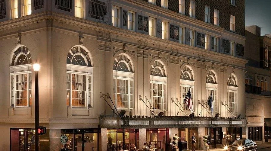 Night view of a historic brick building with bright interior lights, multiple tall arched windows, and several flags displayed outside. A streetlamp illuminates the corner, and cars are parked or driving nearby.