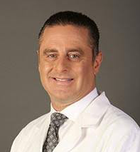 Professional portrait of a male doctor with short dark hair, wearing a white lab coat and a patterned tie, against a plain background.