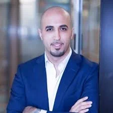 A man with a shaved head and goatee wearing a blue suit and white shirt, standing with arms crossed in an office setting.