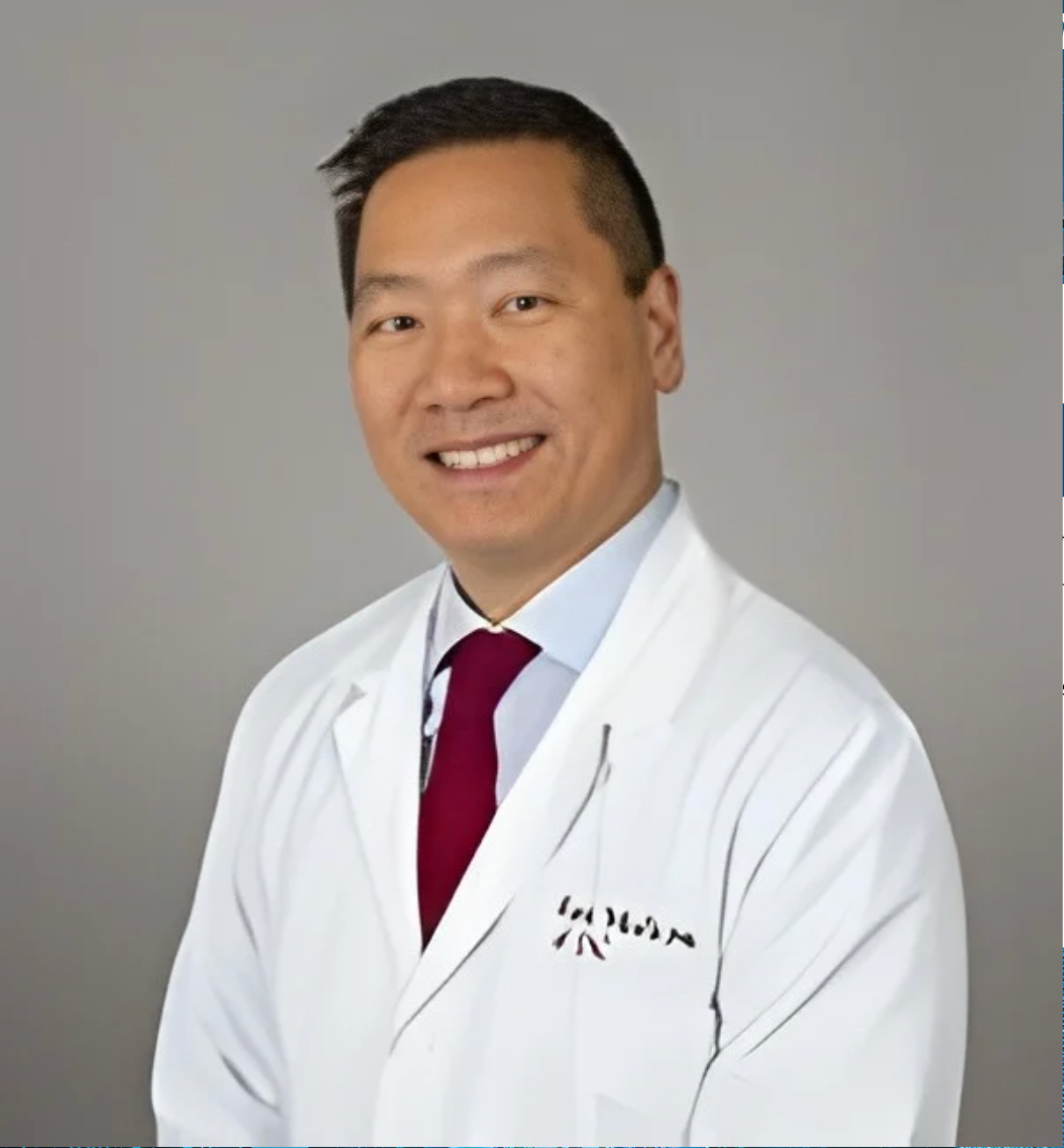 Professional portrait of a smiling male doctor of Asian descent wearing a white lab coat and a maroon tie, against a gray background.