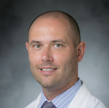 Portrait of a man with a bald head, wearing a white lab coat and a light blue shirt with a tie, against a grey background.