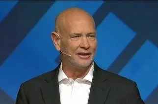 A man with a bald head and a white beard speaking on stage, wearing a black suit and white shirt, with a blue geometric background.