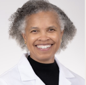A smiling older woman with gray curly hair, wearing a white lab coat and a black shirt, against a light gray background.