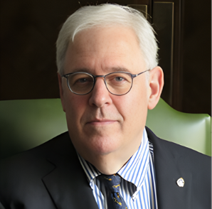 A middle-aged man with white hair wearing glasses, a dark suit, and a blue striped shirt, sitting in front of a dark background.