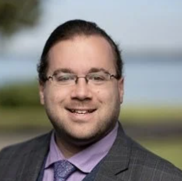A man with glasses, wearing a suit and tie, smiling outdoors with water and trees in the background.