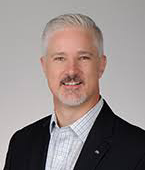 A man with gray hair and a beard smiling, wearing a dark suit jacket and a collared shirt, against a plain gray background.