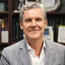 A man in a gray suit smiling in an office setting with shelves and books in the background.