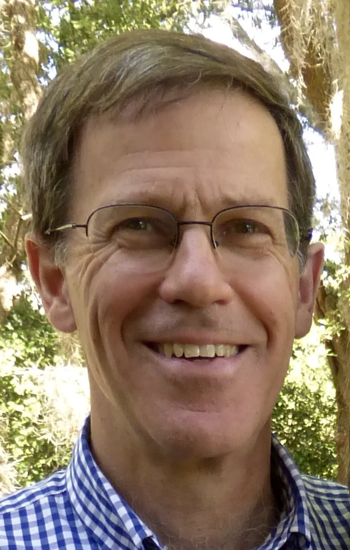 Close-up of a smiling man with glasses and short brown hair, wearing a blue and white checkered shirt outdoors with trees and sunlight in the background.